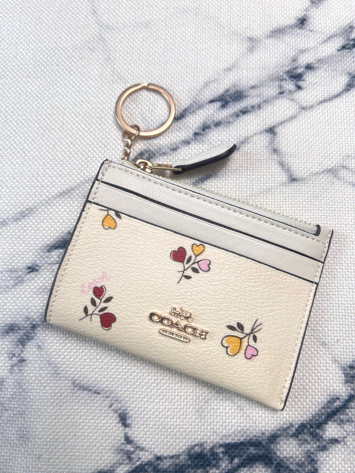 A Coach card holder wallet in off-white pebbled leather with colorful embroidered flowers and the gold-tone Coach logo, shown with a key ring on a white and gray marble-patterned fabric background.