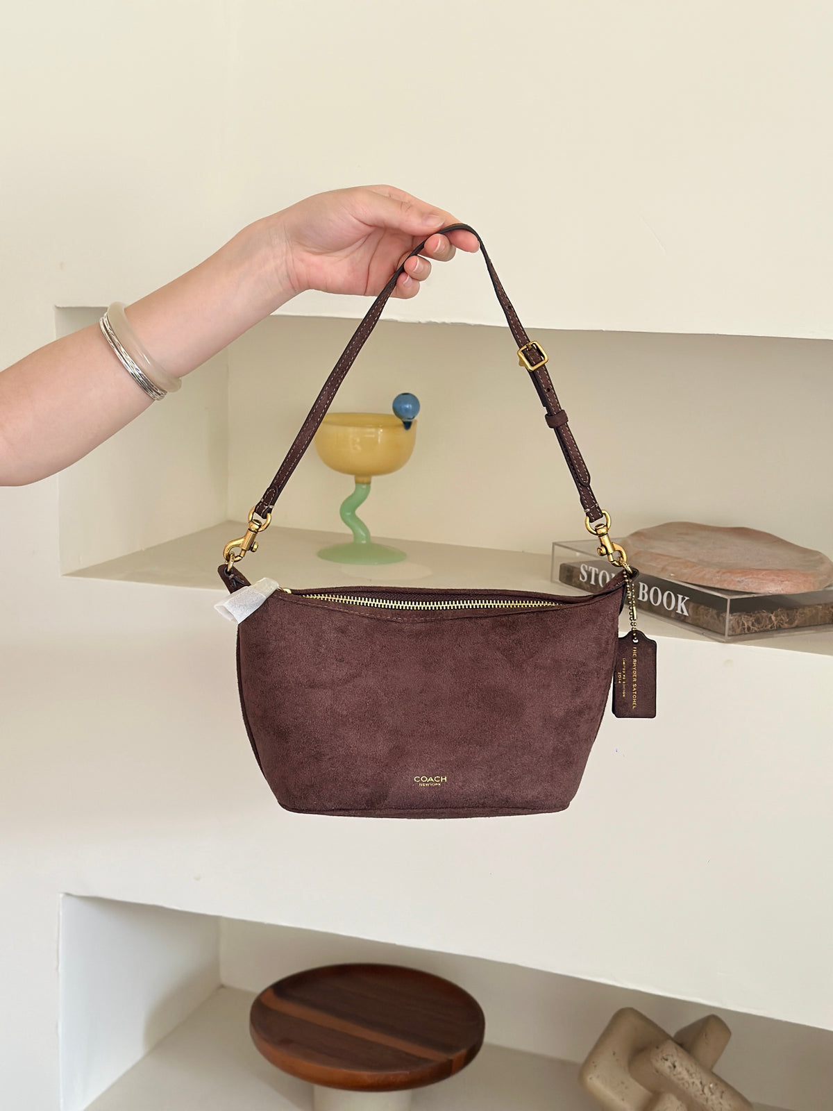 A person's hand holds up a dark brown suede shoulder bag with gold hardware and a zippered top, set against a minimalist cream-colored shelving background.