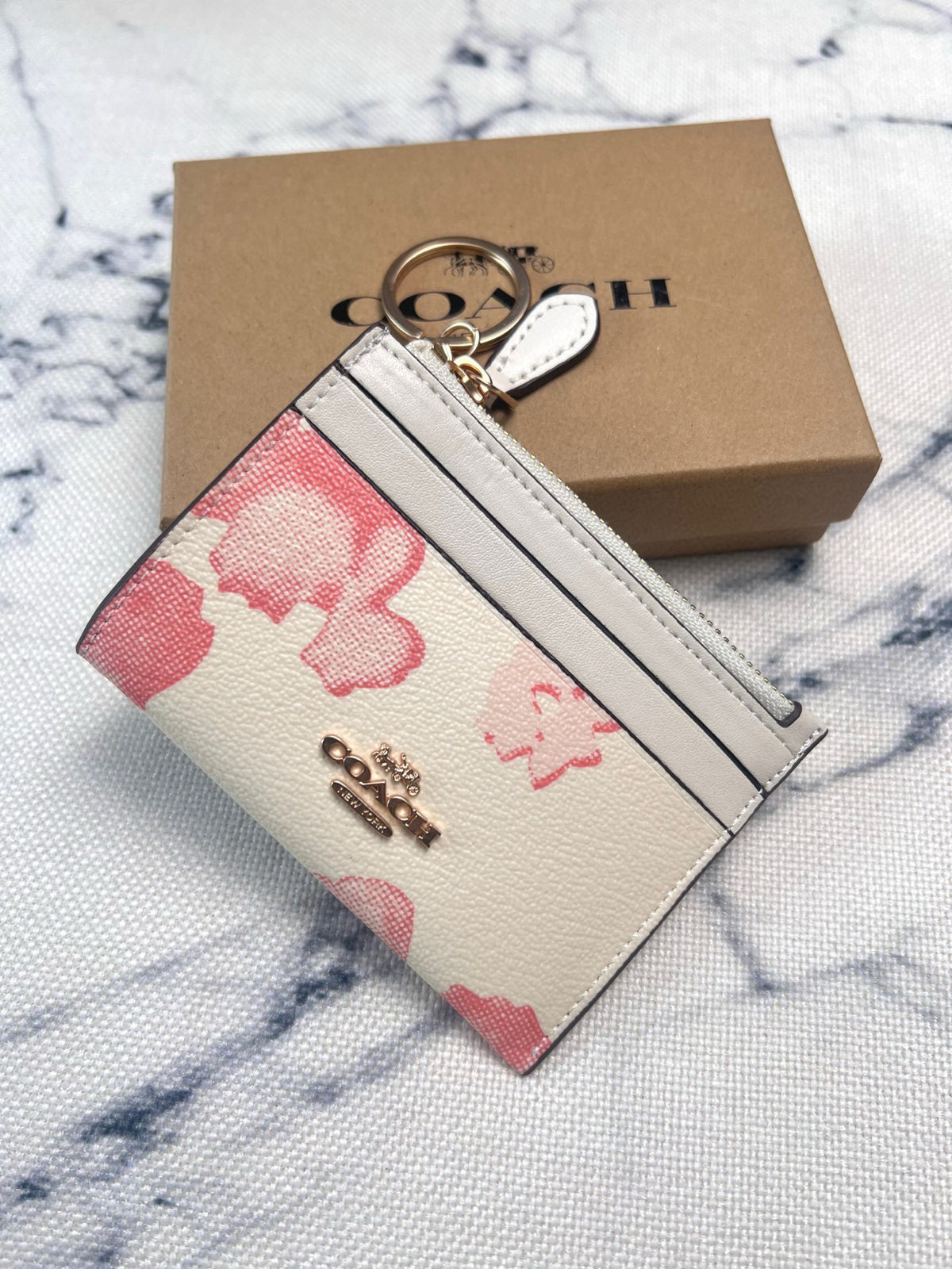 A Coach card holder wallet with a pink floral pattern on a cream textured leather base, featuring a gold-tone metal key ring and a gold-tone Coach logo. It is displayed in front of its brown cardboard packaging box on a white and gray marble-patterned fabric background.
