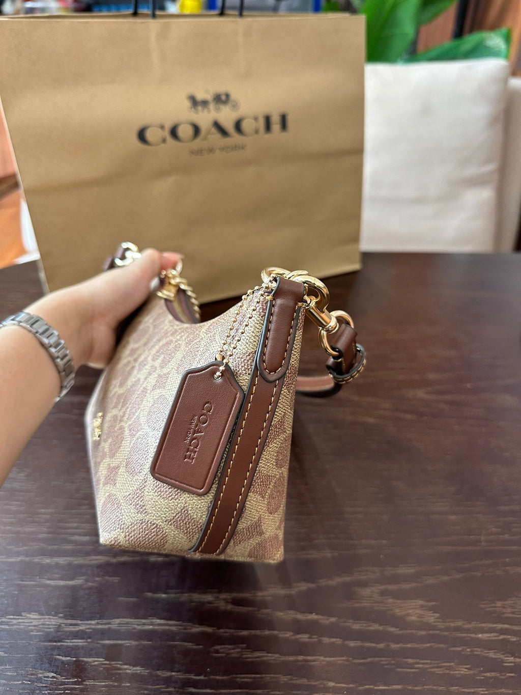 A Coach handbag in signature monogram canvas with brown leather trim and gold-tone hardware, held by a hand in front of a Coach shopping bag on a wooden table.