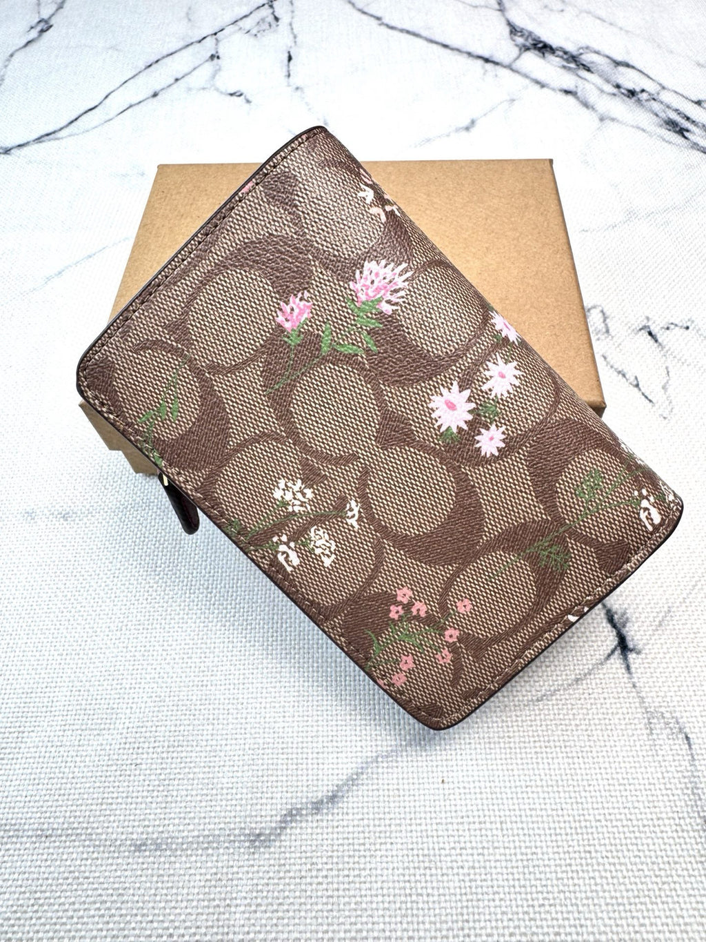 A brown Coach signature canvas wallet with a floral print, resting on a brown cardboard box over a white marble surface.