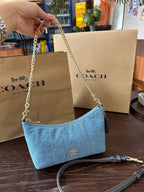 A light blue Coach quilted shoulder bag with a gold-tone chain strap, held by a hand in front of two Coach shopping bags on a dark wooden table.