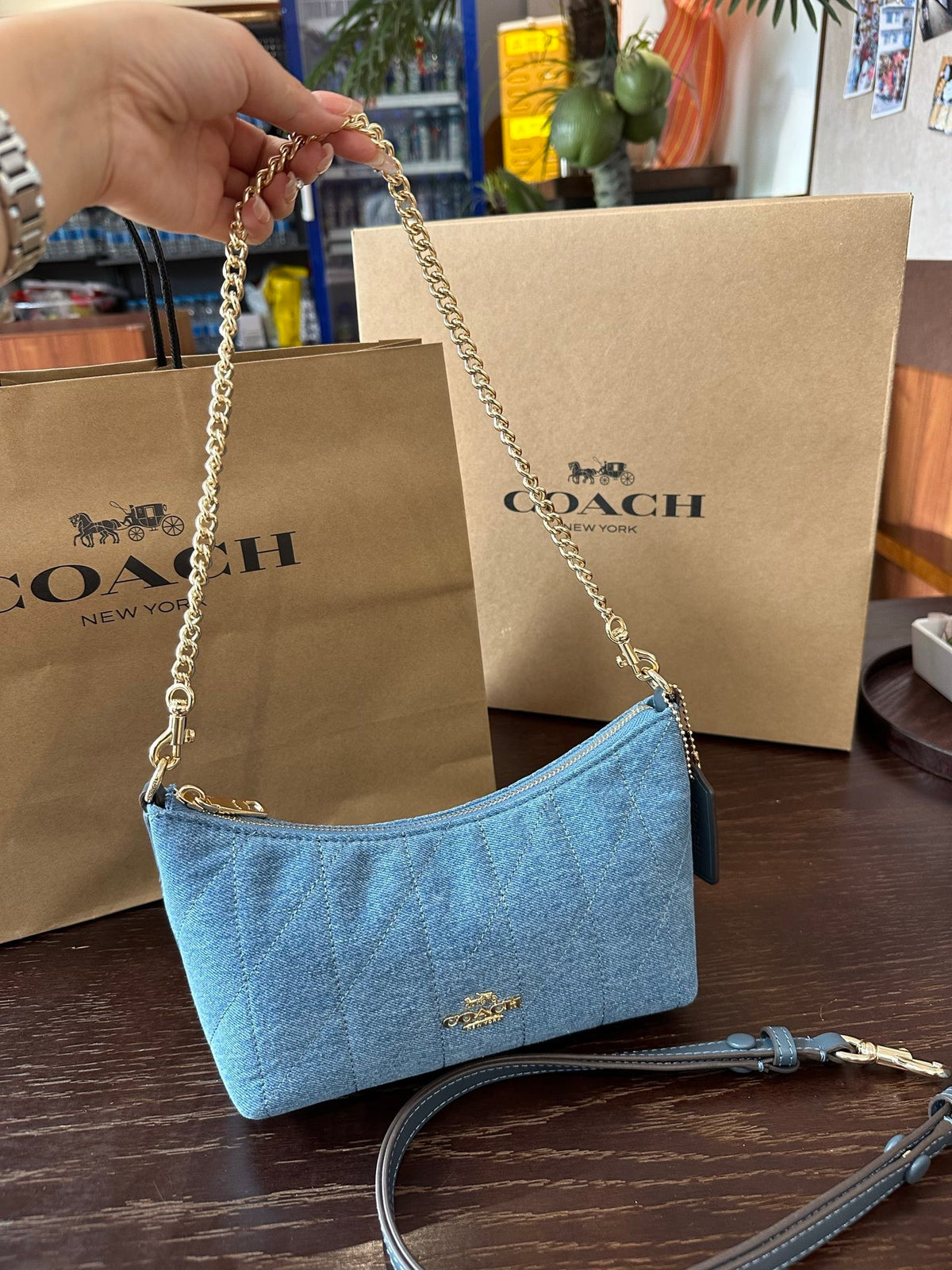 A light blue Coach quilted shoulder bag with a gold-tone chain strap, held by a hand in front of two Coach shopping bags on a dark wooden table.