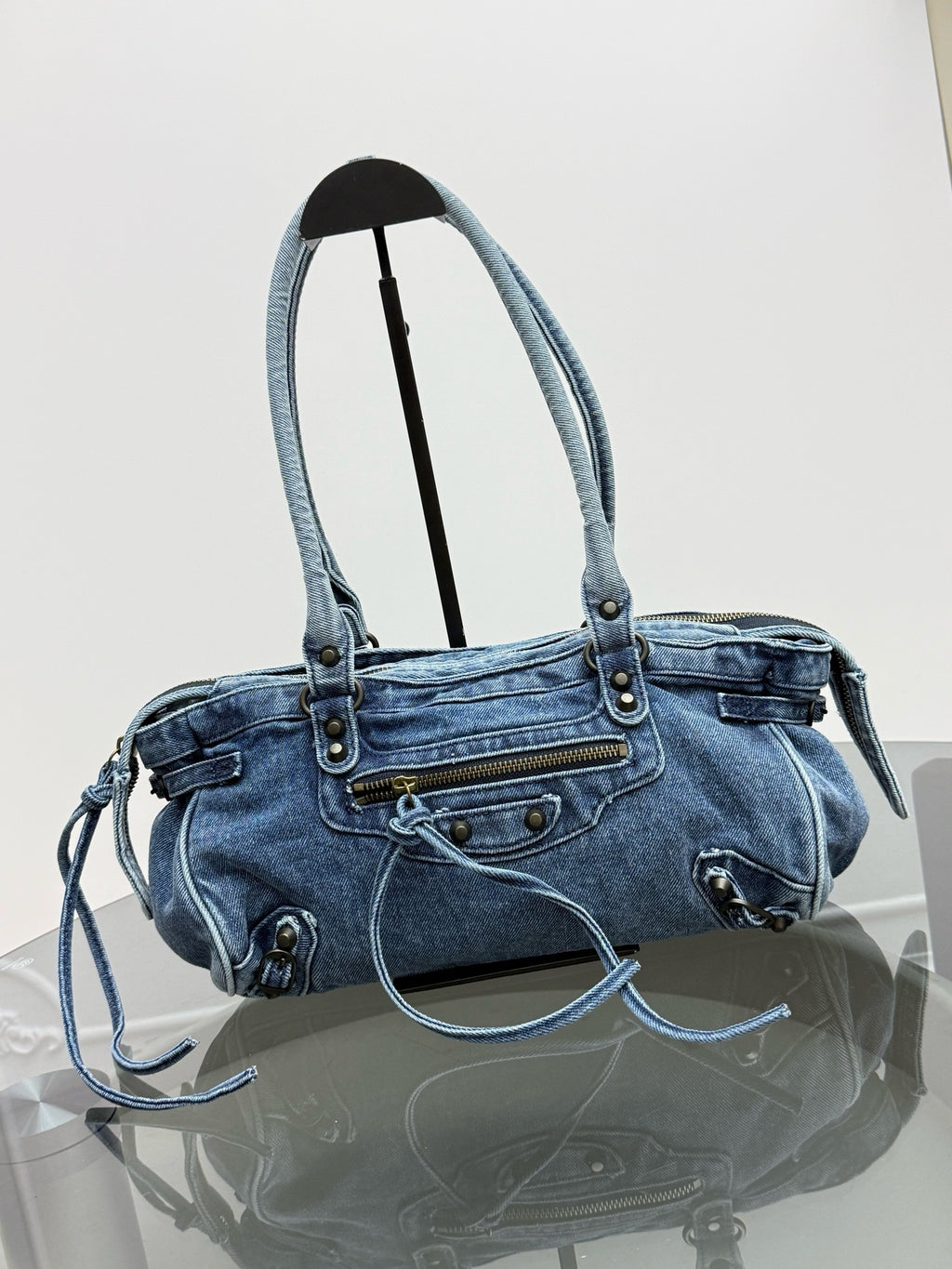 A light blue denim shoulder bag with double top handles, a front zip pocket, and decorative metal studs, displayed on a stand over a reflective surface against a white background.