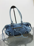 A light blue denim shoulder bag with double top handles, a front zip pocket, and decorative metal studs, displayed on a stand over a reflective surface against a white background.