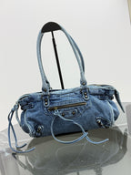 A light blue denim shoulder bag with double top handles, a front zip pocket, and decorative metal studs, displayed on a stand over a reflective surface against a white background.