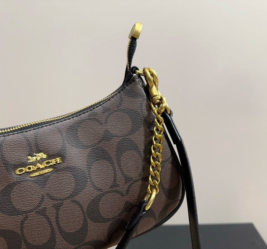 A close-up of a Coach handbag in brown and black monogram canvas with gold-tone hardware, including a chain strap and zipper pull, on a light gray background.