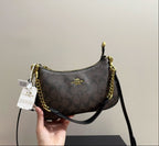 A Coach shoulder bag in brown signature monogram canvas with gold-tone hardware and a black leather strap, held by a hand against a plain beige wall.