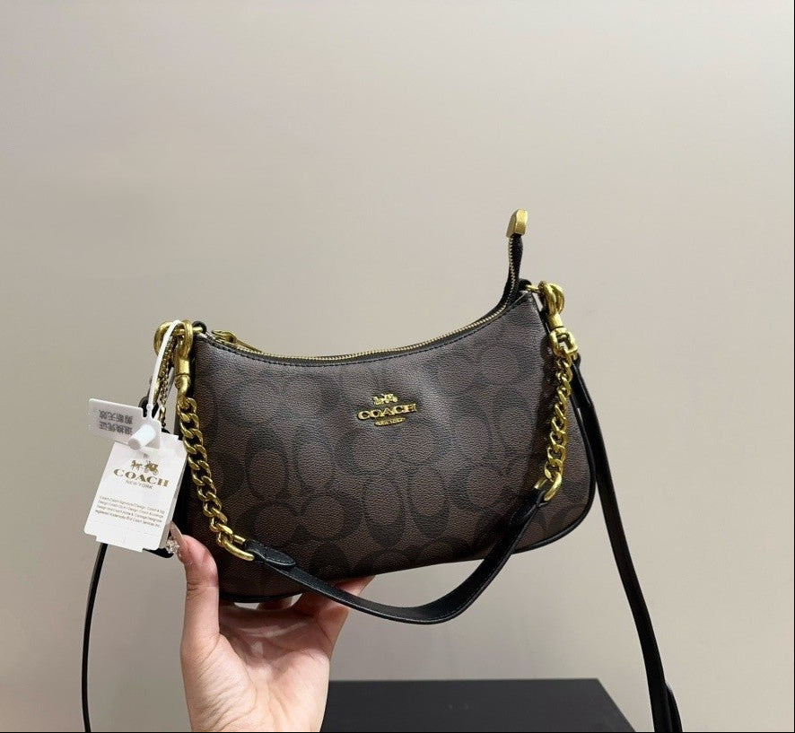 A Coach shoulder bag in brown signature monogram canvas with gold-tone hardware and a black leather strap, held by a hand against a plain beige wall.