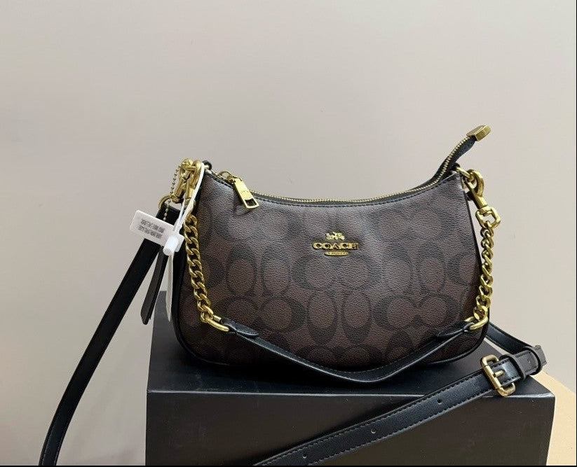 A Coach crossbody bag in the signature brown monogram canvas, displayed on a black box. The bag features gold-tone hardware, a black leather shoulder strap, and a black leather crossbody strap, with a white hangtag attached.