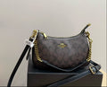 A Coach crossbody bag in the signature brown monogram canvas, displayed on a black box. The bag features gold-tone hardware, a black leather shoulder strap, and a black leather crossbody strap, with a white hangtag attached.