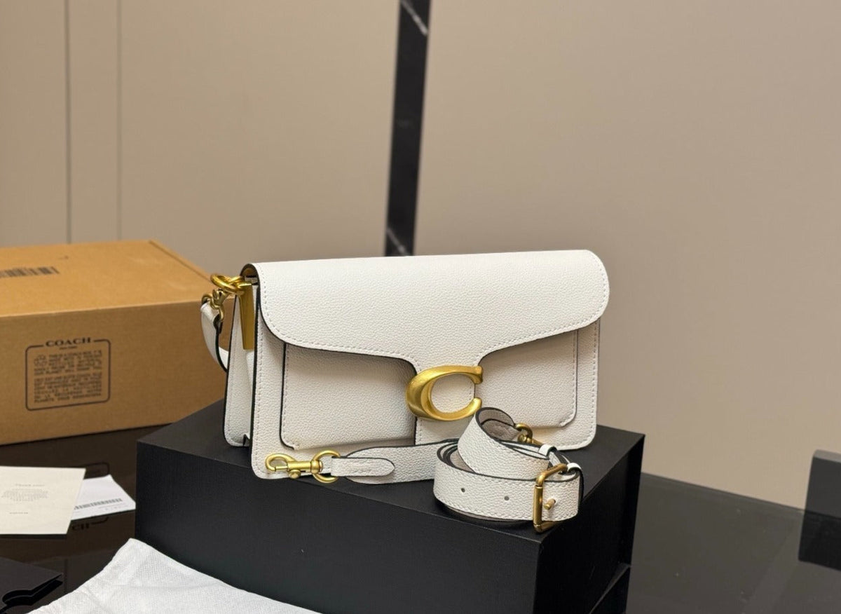A white leather shoulder bag with gold hardware displayed on a black box, alongside its dust bag, papers, and a brown box, set against a beige wall background.