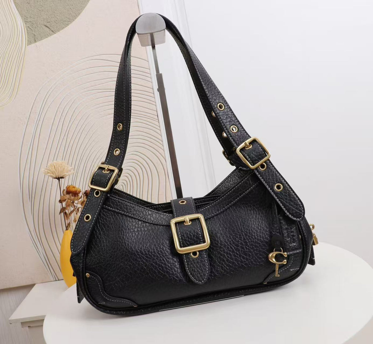 A black pebbled leather shoulder bag with gold-tone hardware, displayed on a stand against a light background with a decorative dried flower prop.
