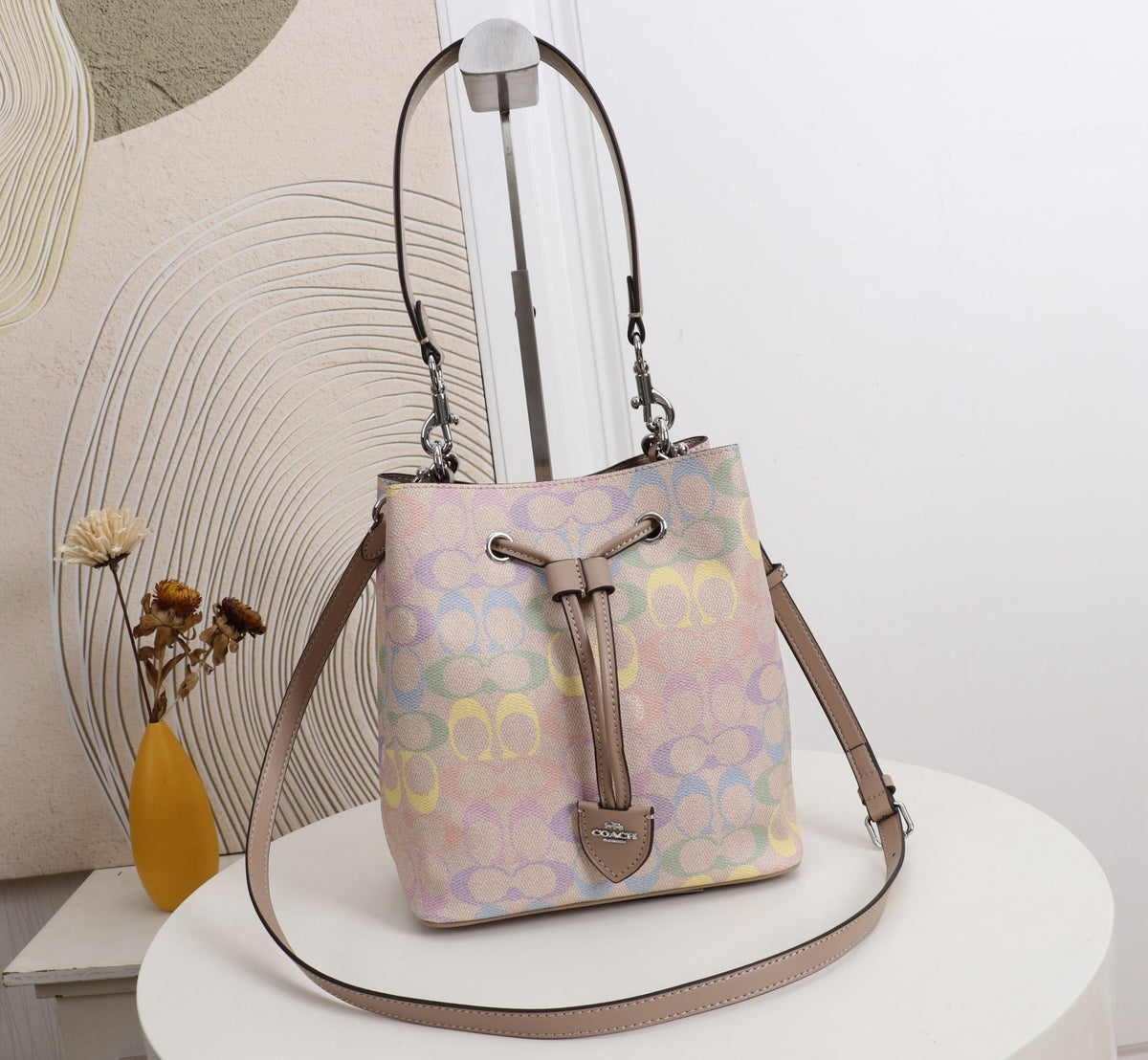 A pastel multicolor bucket bag with a signature monogram pattern, displayed on a white round table. The bag features a drawstring closure, a tan leather strap, and a small gold-tone logo plate at the bottom.