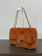 A brown faux fur shoulder bag with a gold-tone chain strap and a small gold turn-lock clasp, displayed on a stand on a dark wooden surface against a light gray background.
