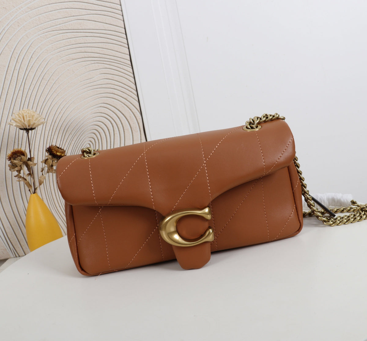 Brown leather handbag with gold chain on a white surface with a decorative wall in the background