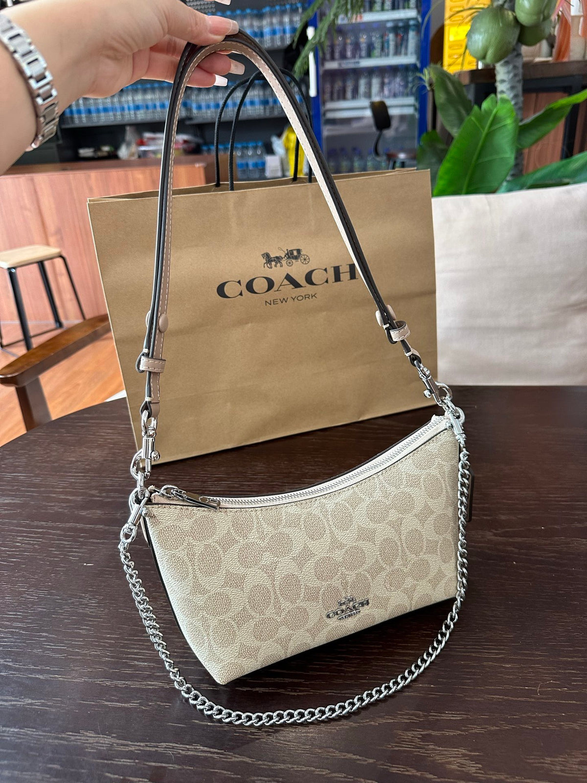 A Coach shoulder bag in beige signature canvas with silver-tone chain and strap details, held by a hand in front of a Coach shopping bag on a dark wooden table.