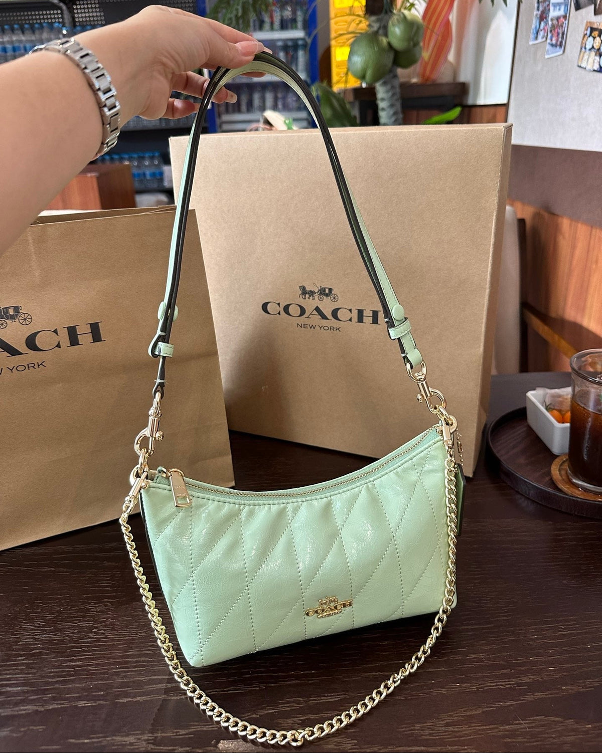 Light green handbag with gold chain held by a hand, with a Coach bag in the background.
