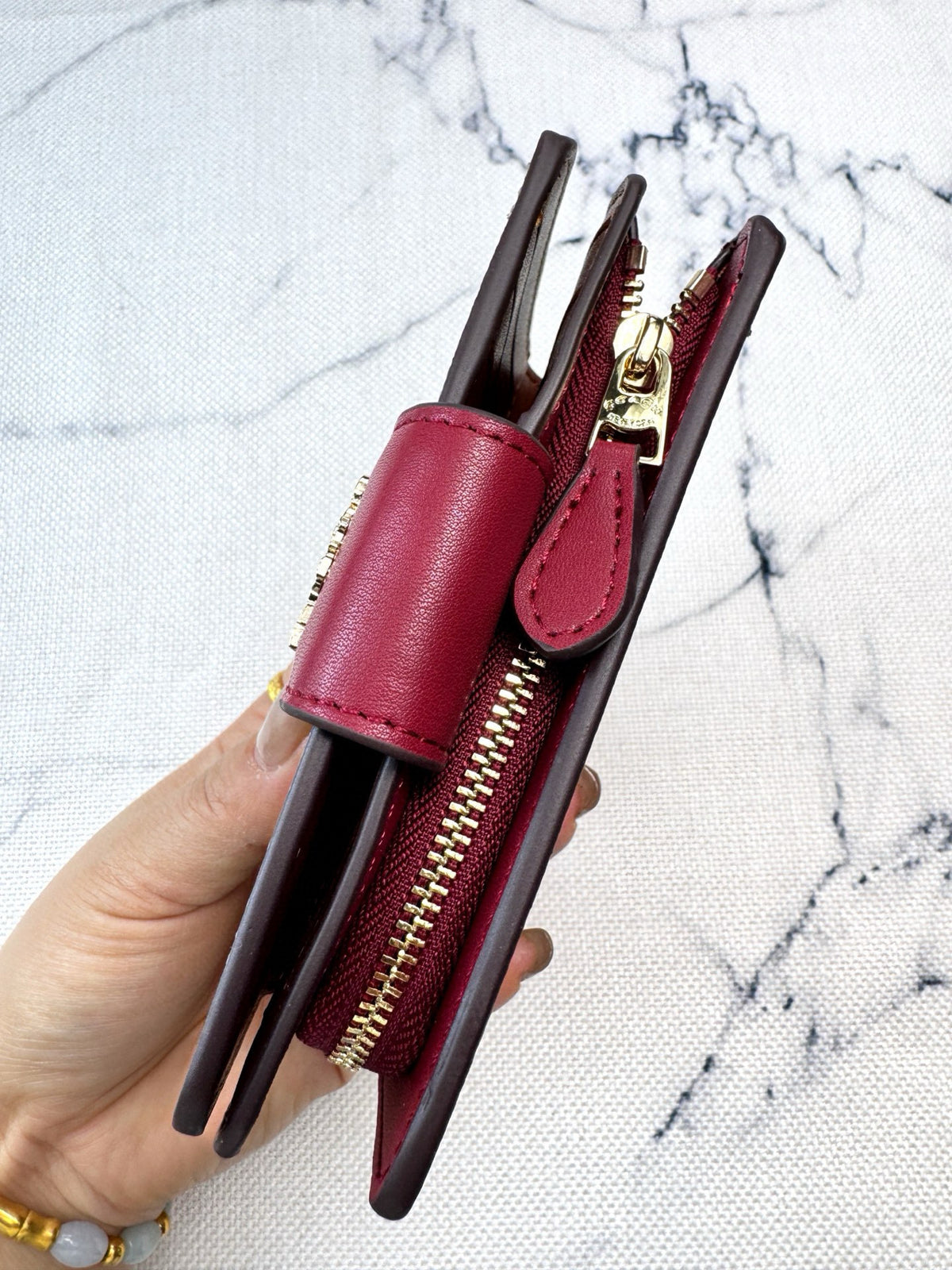 A hand holding a slim burgundy leather wallet with a gold zipper and card slots, set against a white marble background.