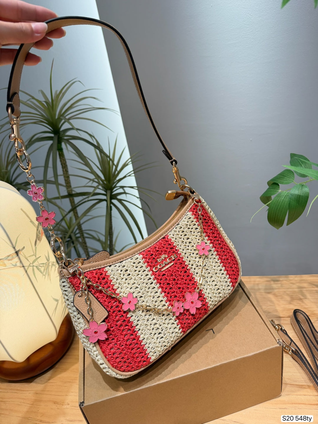 A crocheted shoulder bag with red and cream stripes, decorated with pink flower charms, resting on a brown box on a wooden table with a hand holding its strap.