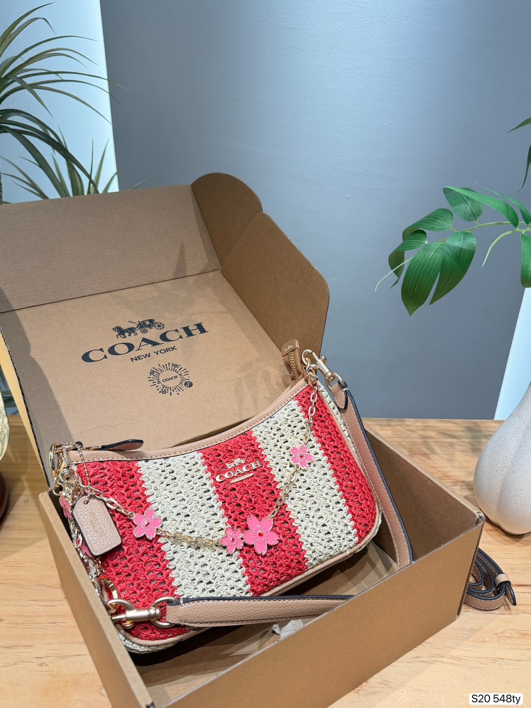 A Coach handbag with red and cream crochet stripes and pink flower charms, displayed inside an open Coach box on a wooden table.