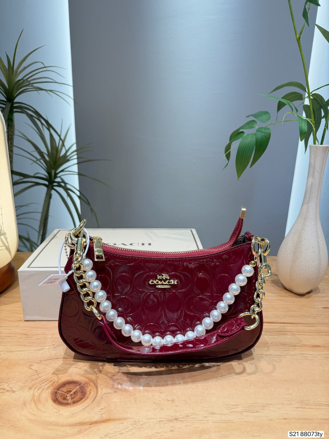 A Coach handbag in deep burgundy patent leather with an embossed monogram pattern, decorated with a white pearl strap and gold-tone chain links, displayed on a wooden table next to a white Coach box.