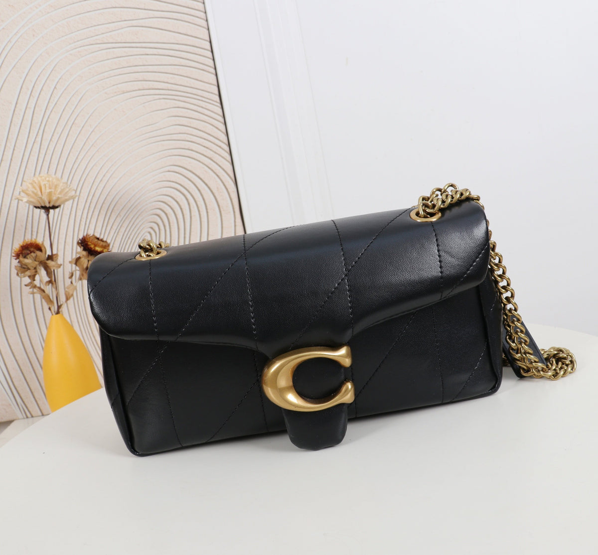 Black quilted handbag with gold chain and clasp on a white surface with a decorative wall in the background.