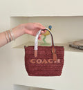 small woven Coach tote bag with a burgundy body, tan leather trim, and a white protective wrap on the handle, set against a cream-colored shelving background.