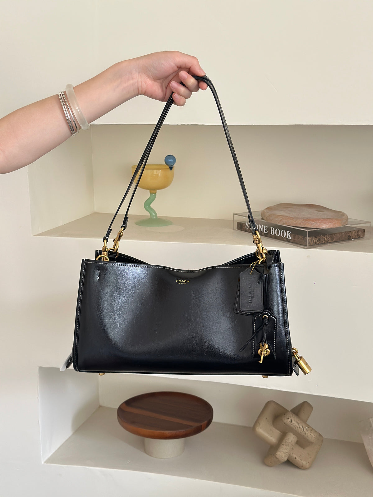 A black leather shoulder bag with gold-tone hardware and a small gold logo on the front, held by a person's hand against a beige shelving background.