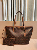 A brown monogram tote bag with tan leather handles and trim, shown with a matching zip pouch on a light wood surface in front of beige curtains.