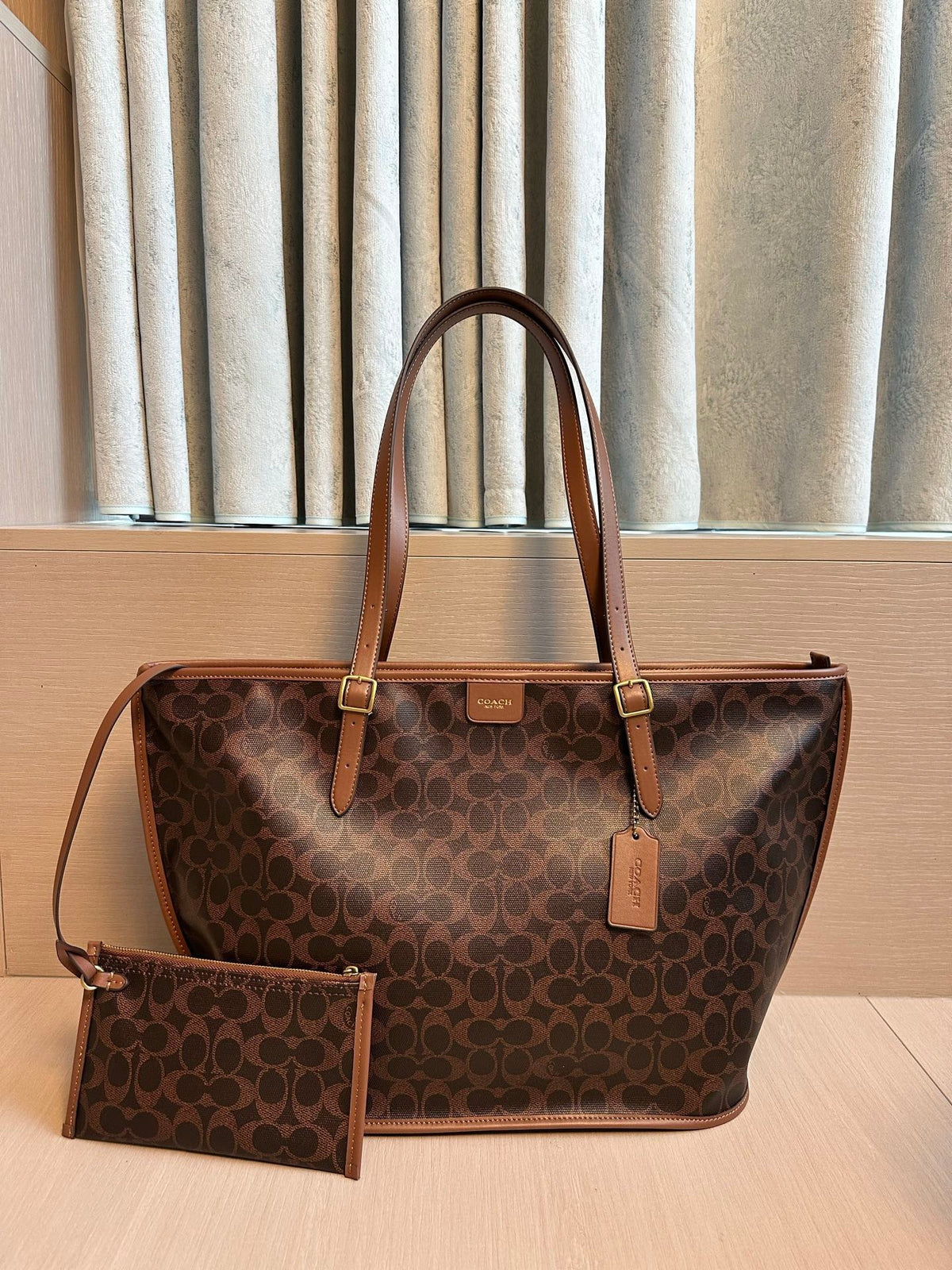 A brown monogram tote bag with tan leather handles and trim, shown with a matching zip pouch on a light wood surface in front of beige curtains.