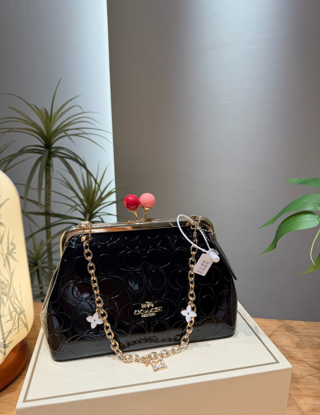 A Coach black patent leather kiss-lock handbag with a gold-tone chain strap and decorative flower charms, displayed on a cream-colored box with a neutral gray background and indoor plants.
