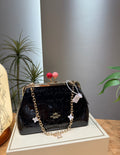 A Coach black patent leather kiss-lock handbag with a gold-tone chain strap and decorative flower charms, displayed on a cream-colored box with a neutral gray background and indoor plants.