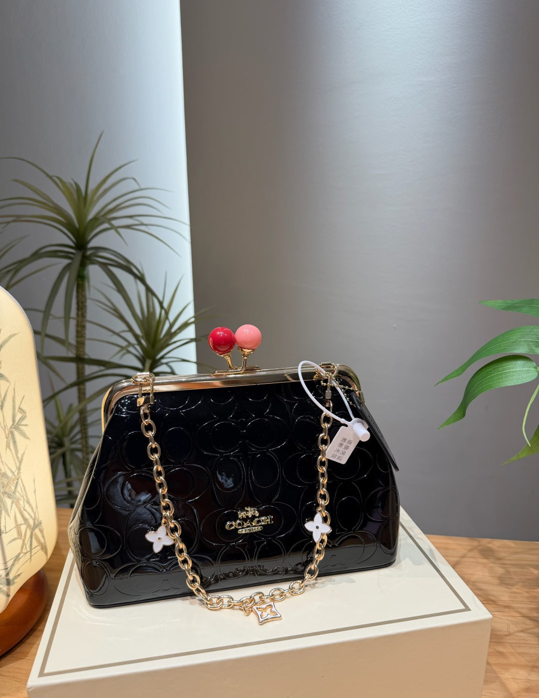 A Coach black patent leather kiss-lock handbag with a gold-tone chain strap and decorative flower charms, displayed on a cream-colored box with a neutral gray background and indoor plants.