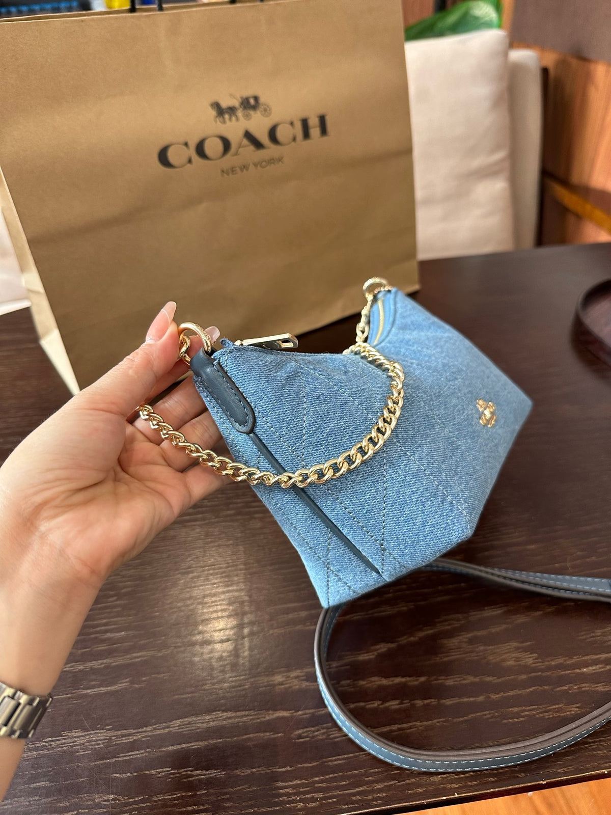 A light blue Coach shoulder bag with gold hardware and a chain strap, held by a hand in front of a brown Coach shopping bag on a dark wooden table.