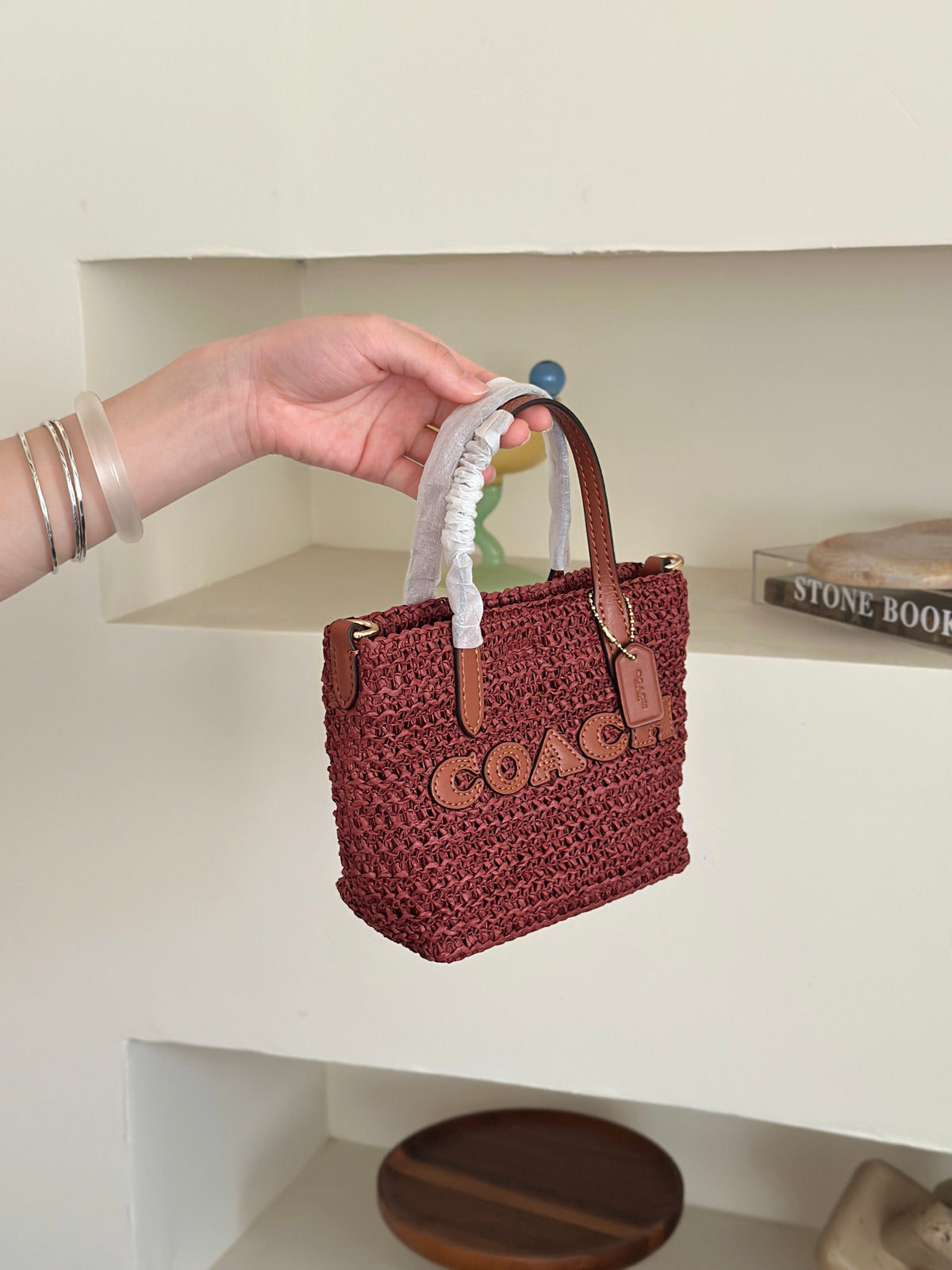 small Coach woven straw tote bag with brown leather handles and a white fabric wristlet strap, set against a minimalist cream-colored shelf background.