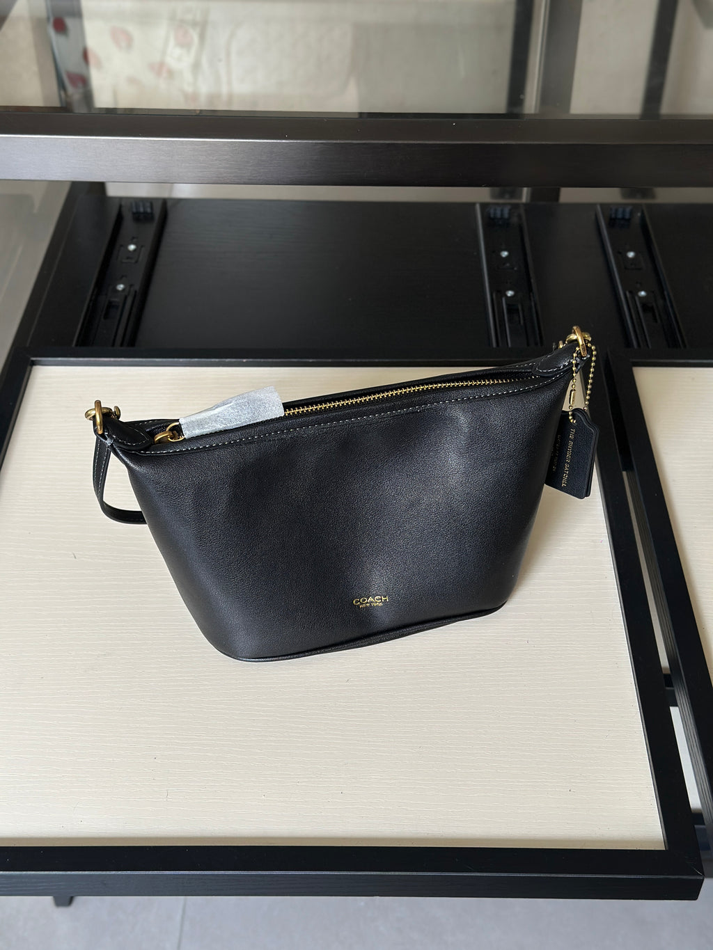 A black leather shoulder bag with gold-tone hardware and a small gold logo, displayed on a cream-colored shelf of a black metal display stand.
