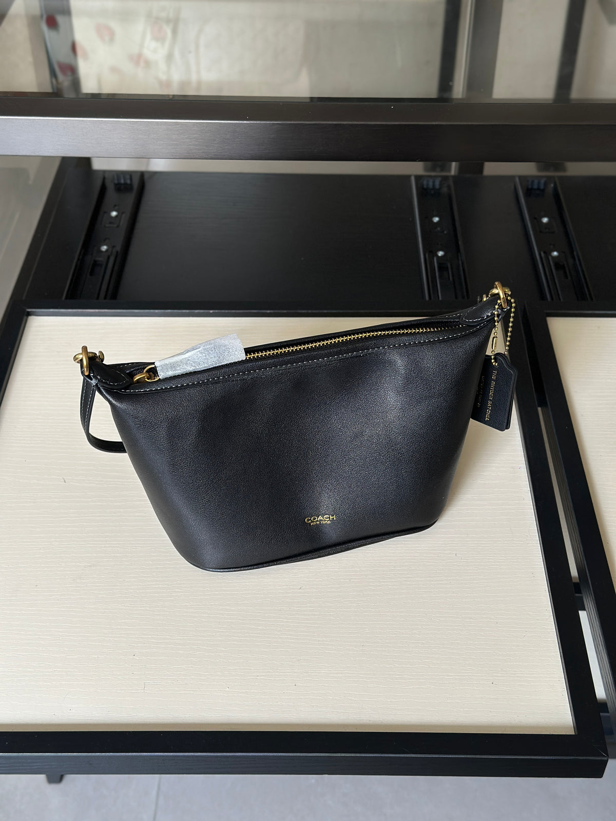 A black leather shoulder bag with gold-tone hardware and a small gold logo, displayed on a cream-colored shelf of a black metal display stand.