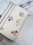 A close-up of a Coach wallet in textured cream-colored leather with colorful embroidered floral motifs and a gold-tone Coach logo on the front, resting on a light marble-patterned surface.