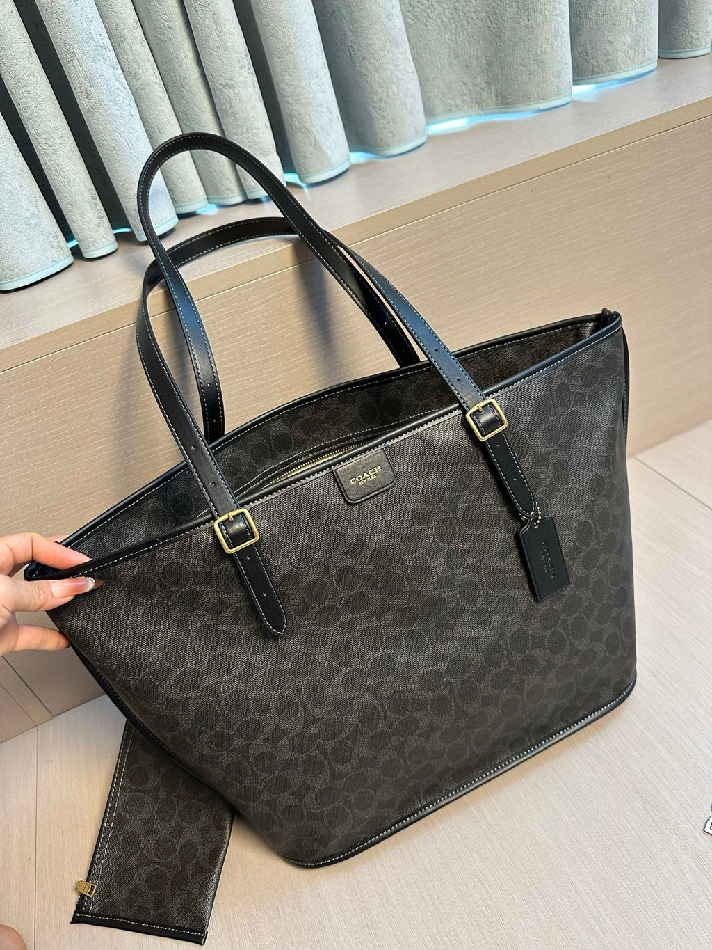 A Coach tote bag in dark brown and black signature monogram canvas, held by a hand on a light wood floor in front of a window with gray curtains.