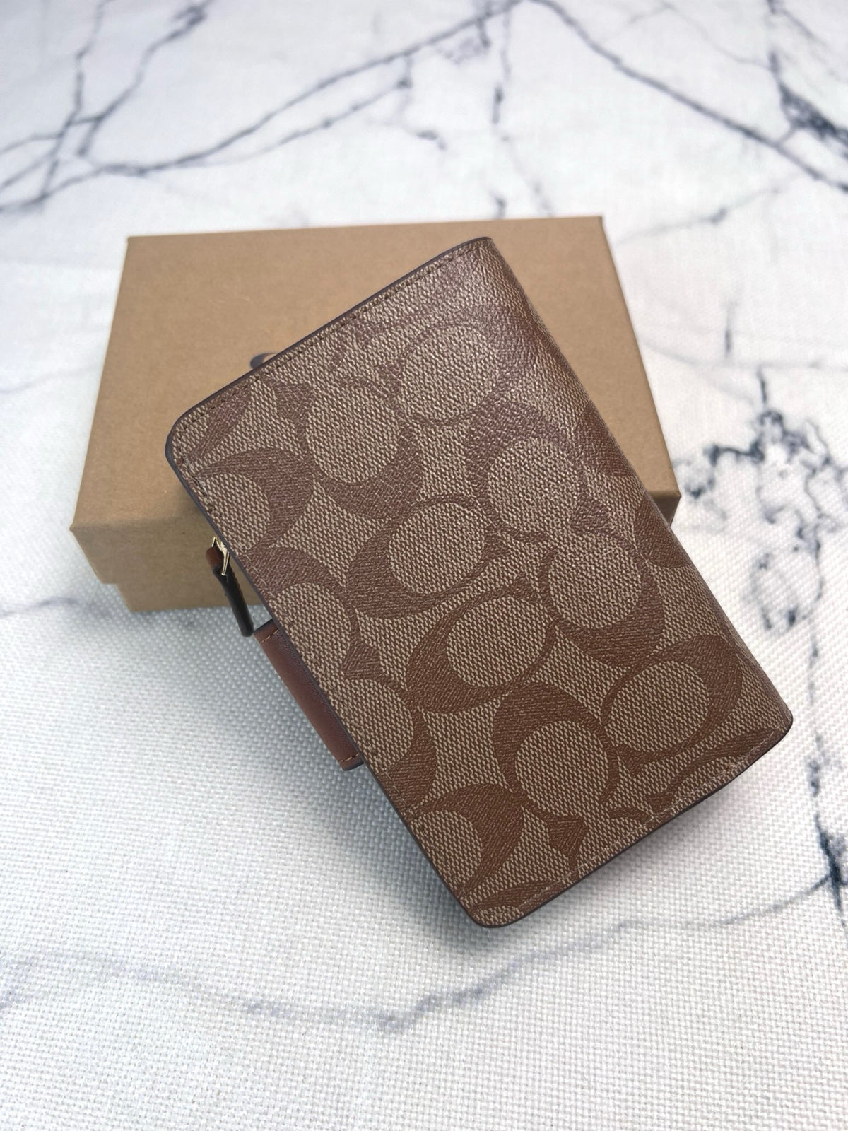 A brown and tan monogram wallet or card holder with a small side zipper, placed diagonally on top of a plain brown box over a white marble-like surface.