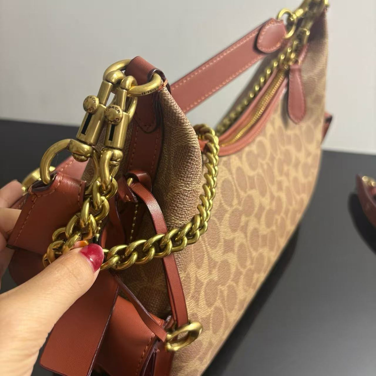 A close-up of a beige and brown monogram handbag with pink leather trim and gold-tone hardware, held by a hand with pink nail polish.