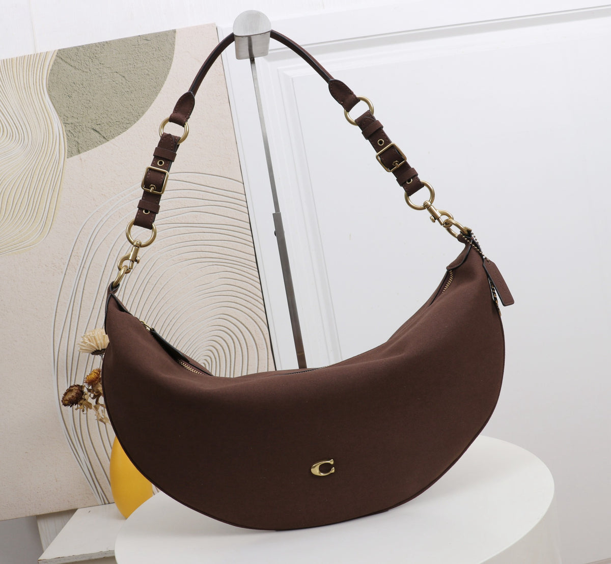 A brown crescent-shaped shoulder bag with a gold-tone C logo, displayed on a white pedestal. The bag features a matching brown leather shoulder strap with gold-tone hardware and a small decorative tassel.