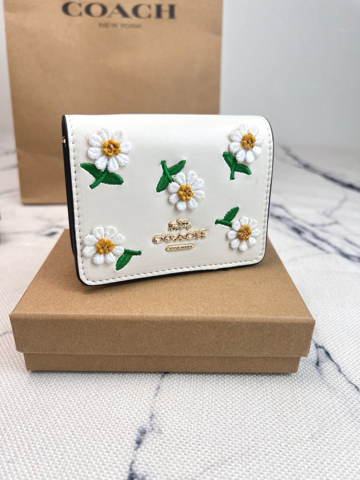 A Coach white leather wallet decorated with embroidered daisy flowers, displayed on a brown cardboard box with a Coach bag in the background.