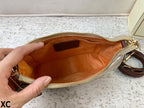 A close-up of a hand holding open a beige and brown clutch or pouch, showing a tan interior lining and gold-tone zipper. A matching brown strap is visible on the right, and the pouch is photographed on a light textured surface.