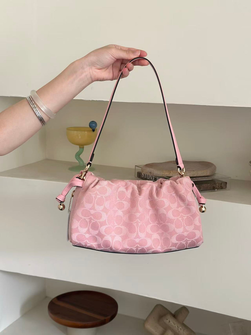 A pink Coach handbag with a signature monogram pattern is held by a person's hand in front of white shelves with decorative items.