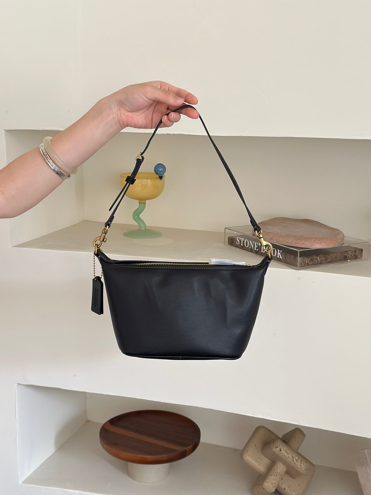 A hand holds up a black leather shoulder bag with a gold zipper and gold hardware, set against a minimalist white shelving background with decorative objects.