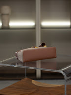 A structured rectangular handbag in dusty pink leather with gold-tone hardware and a small checkered fabric accent panel, displayed on a transparent glass table in a modern interior setting.
