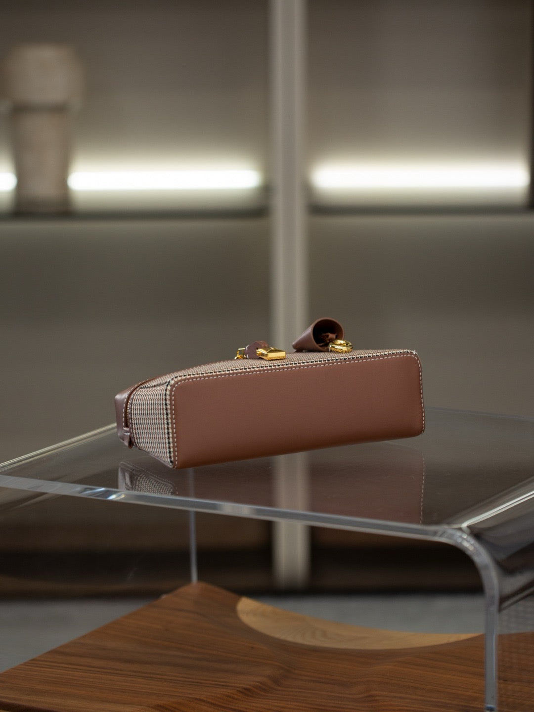 A structured rectangular handbag in dusty pink leather with gold-tone hardware and a small checkered fabric accent panel, displayed on a transparent glass table in a modern interior setting.