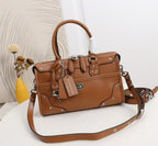A brown leather handbag with a top handle, front flap pockets, silver-tone studs and hardware, and a detachable shoulder strap, displayed on a white surface with a decorative vase in the background.